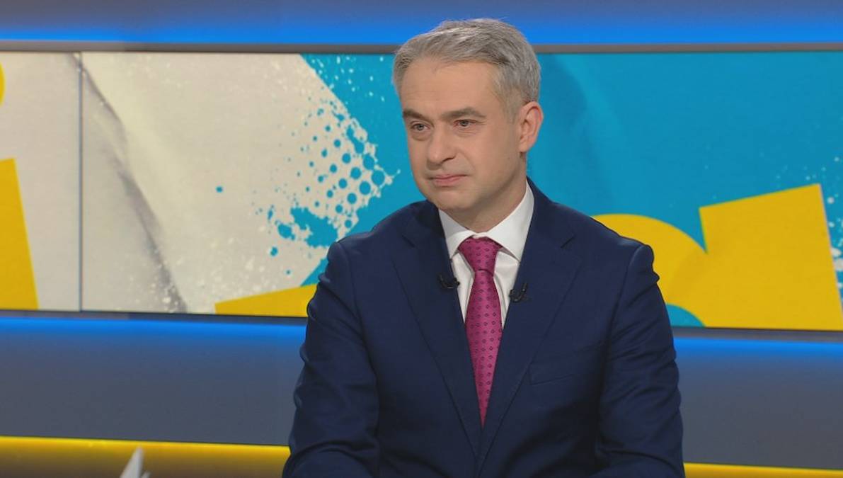 Mężczyzna w granatowym garniturze i różowym krawacie siedzi w studiu telewizyjnym.