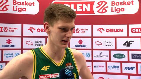 Młody mężczyzna w zielonej koszulce do koszyk&oacute;wki stoi przed ścianą z logo "Basket Liga" i sponsor&oacute;w.