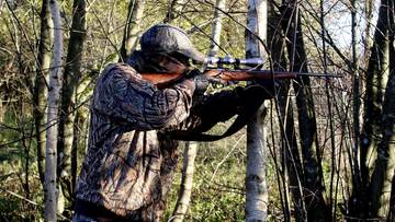 Myśliwy w ubraniu maskującym celuje z karabinu myśliwskiego z lunetą, stojąc w lesie pośród drzew.