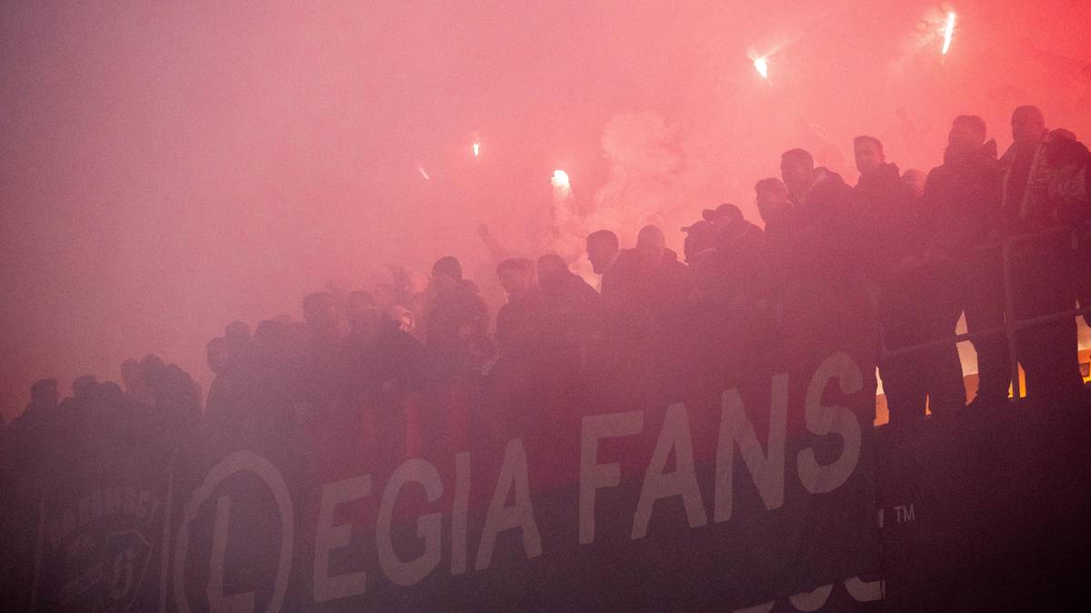 Tłum kibiców Legii Warszawa w dymie z rac, widoczny baner "LEGIA FANS"