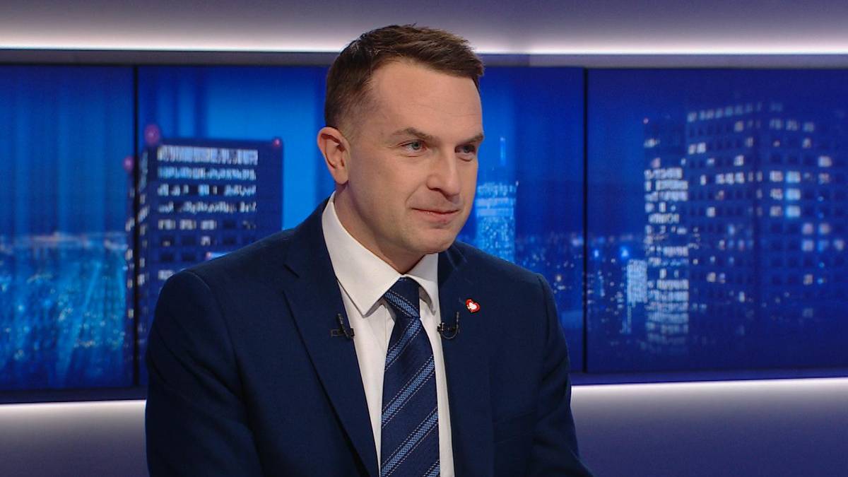 Mężczyzna w garniturze i krawacie siedzi i m&oacute;wi, w tle widać niebieskie ekrany z widokiem miasta.