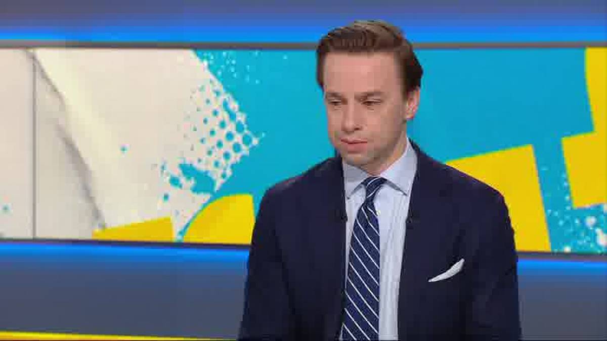 Mężczyzna w garniturze i krawacie siedzi przed kolorowym, geometrycznym tłem.