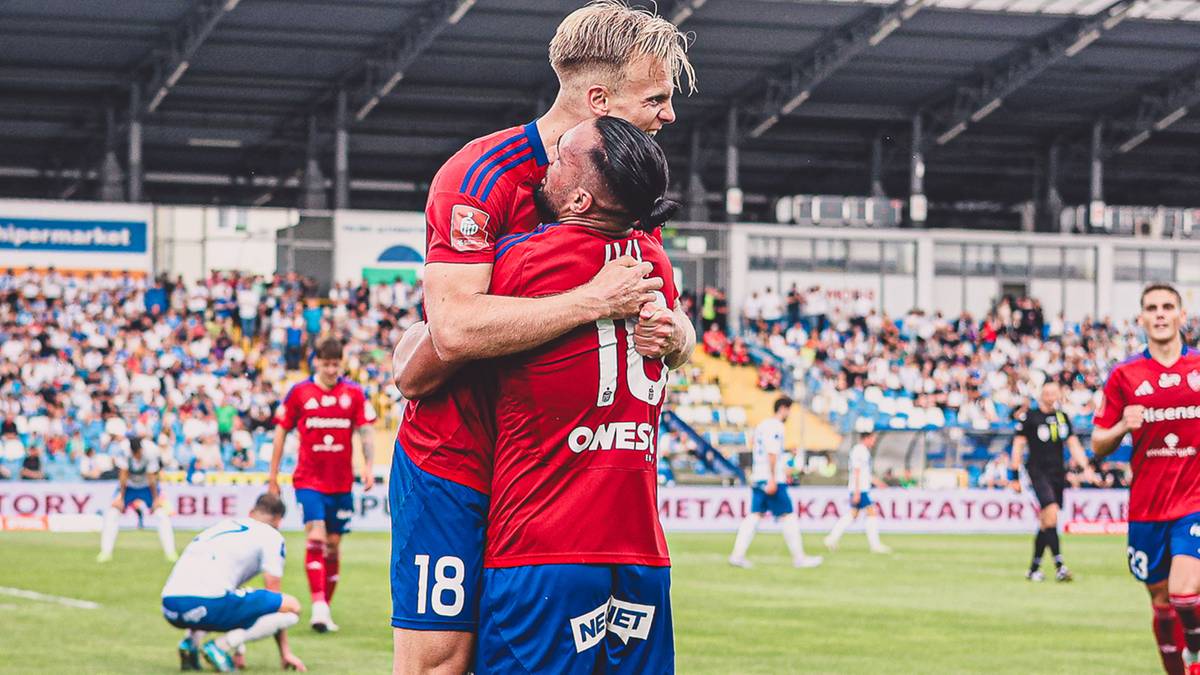 Dw&oacute;ch piłkarzy w czerwonych strojach świętuje na boisku piłkarskim.