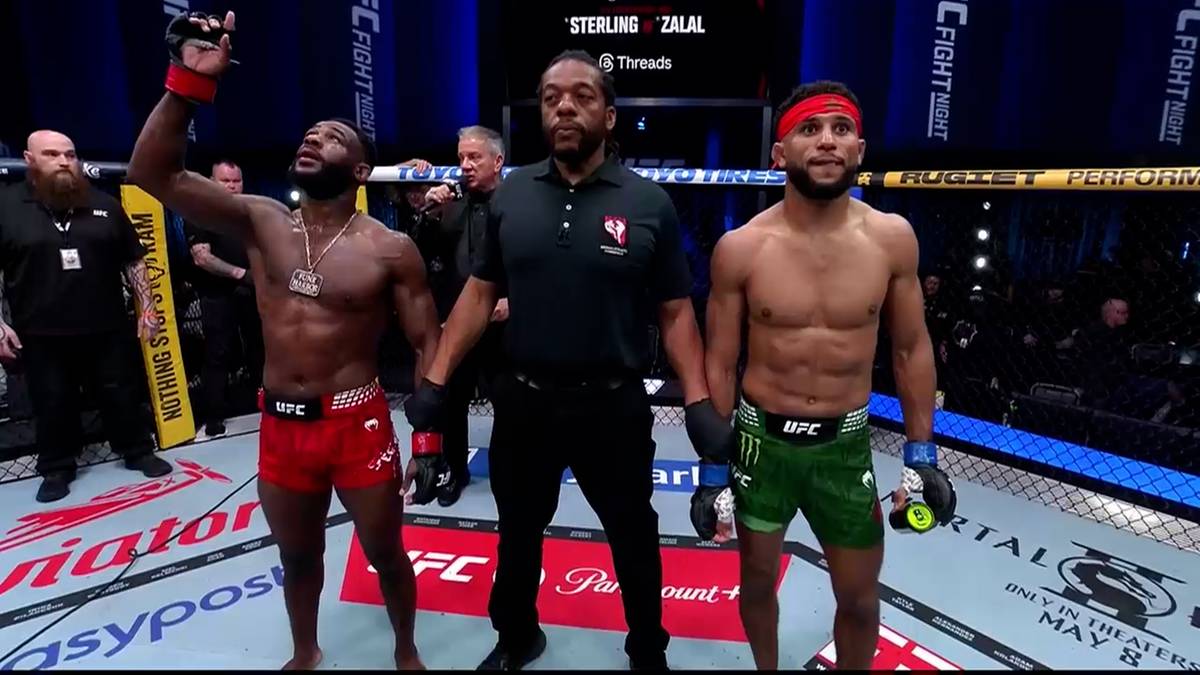 Dwóch zawodników MMA i sędzia stoją na środku klatki po walce. Zawodnik po lewej, w czerwonych spodenkach, unosi rękę w geście zwycięstwa. Zawodnik po prawej, w zielonych spodenkach, stoi ze spuszczoną głową.