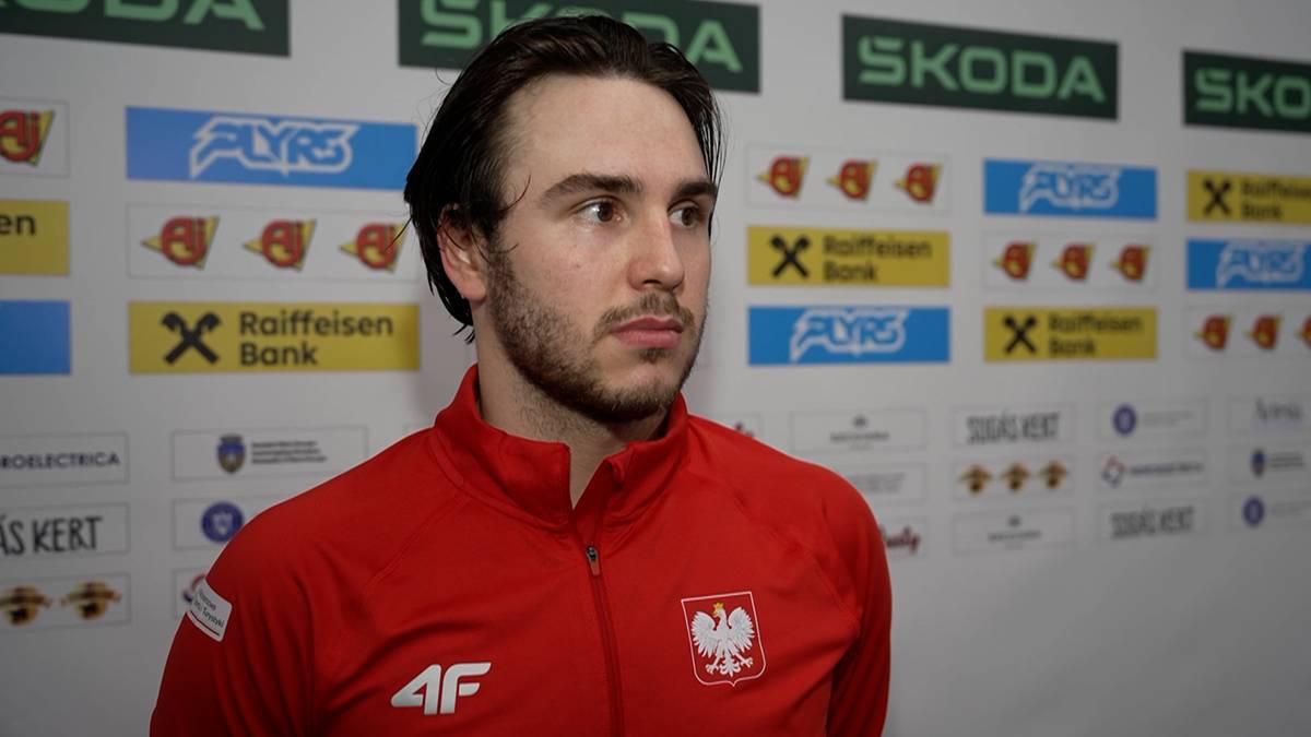 Portret mężczyzny z brodą, ubranego w czerwoną bluzę sportową z logo reprezentacji Polski. Za nim widoczne logo sponsorów i nazwiska.