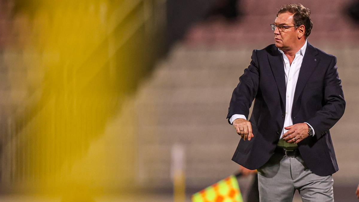 Trener piłkarski w garniturze stojący na boisku i wskazujący ręką.