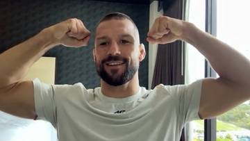 Mateusz Gamrot, polski zawodnik MMA, uśmiecha się i pokazuje swoje bicepsy na tle okna.