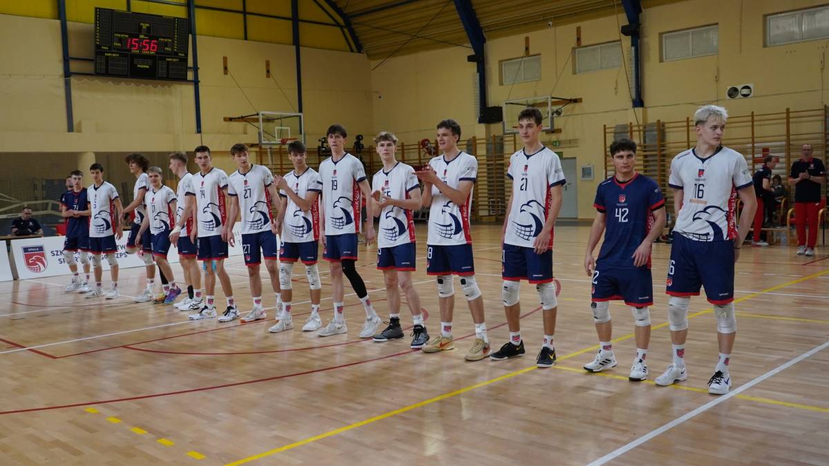 Grupa młodych mężczyzn w strojach sportowych stoi w szeregu na parkiecie sali gimnastycznej.