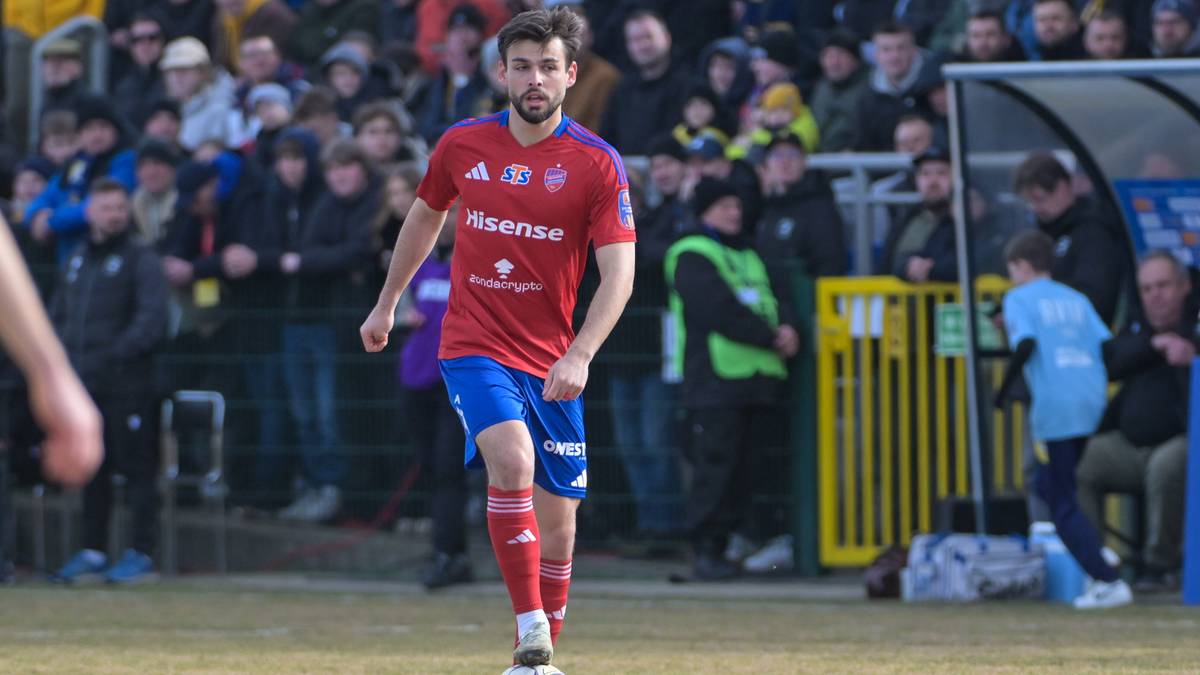 Piłkarz w czerwonej koszulce i niebieskich spodenkach stoi na boisku z piłką przy nodze, na trybunach widoczni kibice.