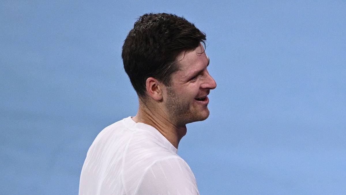 Dwóch tenisistów w białych koszulkach i czerwonych spodenkach ściska sobie dłonie na tle kortu tenisowego.