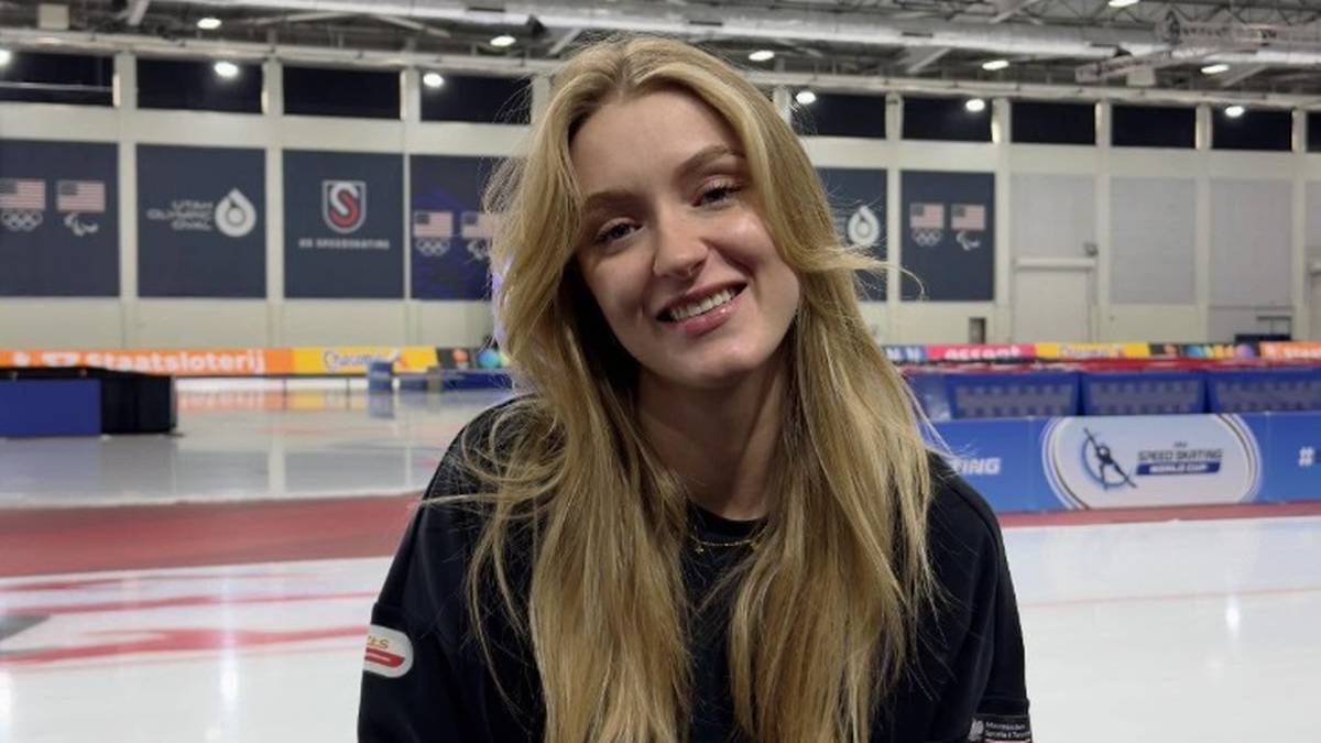 Portret uśmiechniętej młodej kobiety z długimi blond włosami, siedzącej w obiekcie sportowym z lodowiskiem w tle.