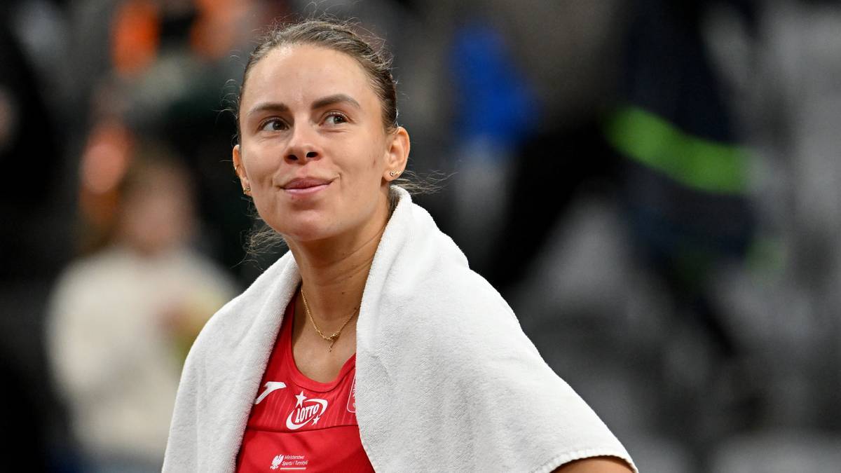 Magda Linette w czerwonej bluzce tenisowej z białym ręcznikiem na ramionach, uśmiechnięta, patrząca w bok.