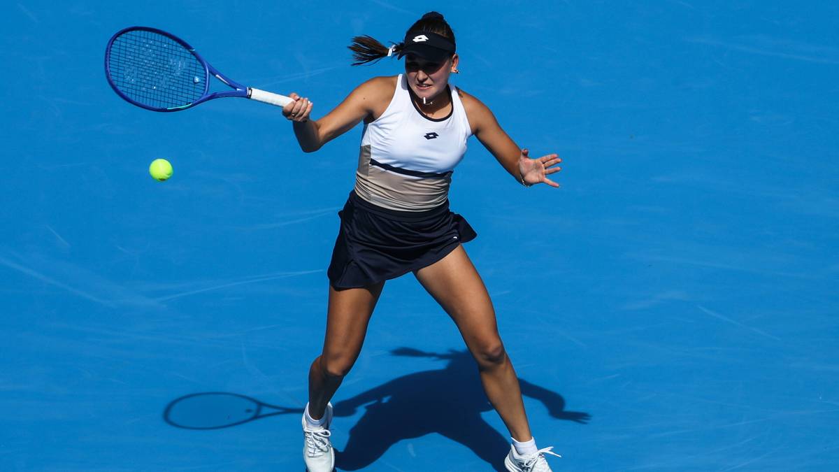 Kobieta w białej koszulce i czarnej spódniczce tenisowej uderza rakietą piłkę tenisową na niebieskim korcie.