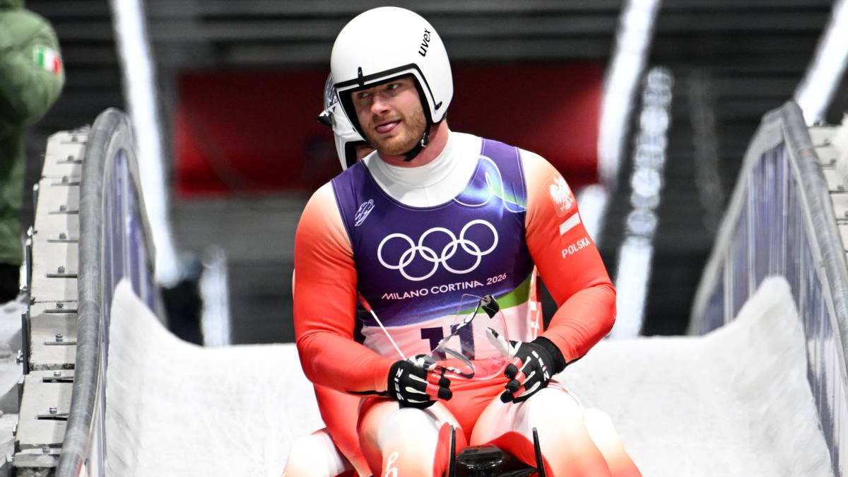 Saneczkarz w kasku i kombinezonie olimpijskim siedzi na sankach na torze lodowym.