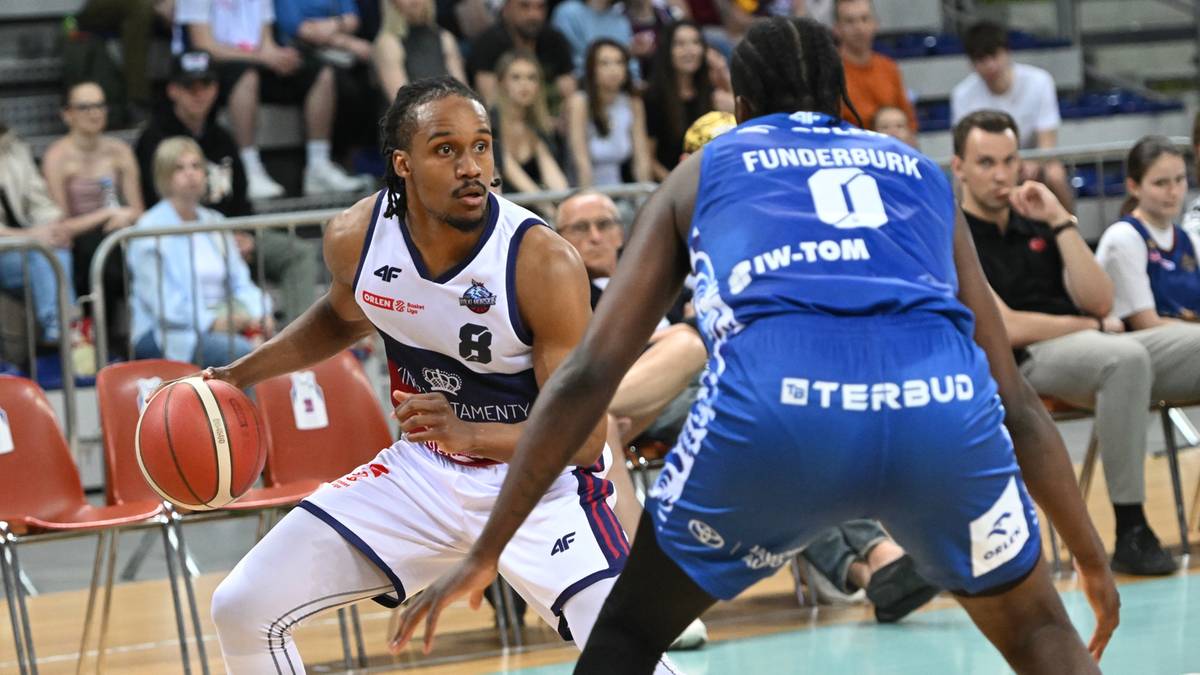 Dwóch zawodników koszykówki na boisku, jeden w białym stroju z numerem 8 próbuje zdobyć punkt, drugi w niebieskim stroju broni. W tle siedzą kibice.