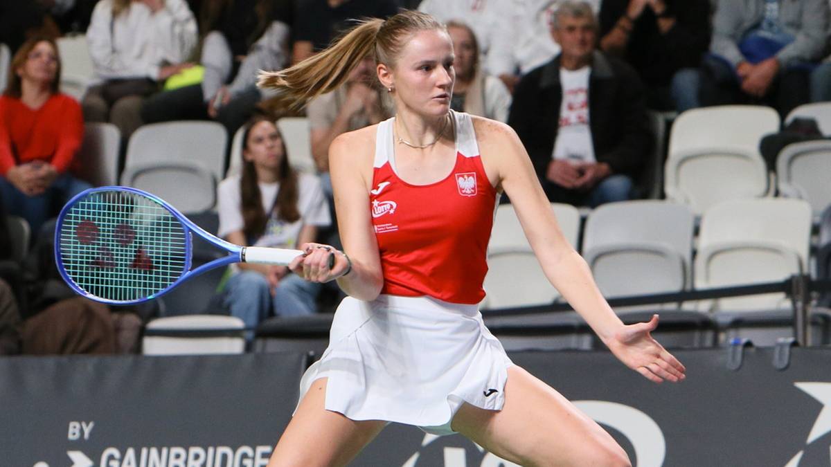 Tenisistka wykonuje zamach rakietą na korcie tenisowym, w białej sp&oacute;dniczce i czerwonym topie.