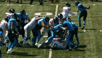 Gracze drużyny w niebieskich strojach próbują zatrzymać gracza drużyny w białych strojach, który przewrócił się na ziemię z piłką.