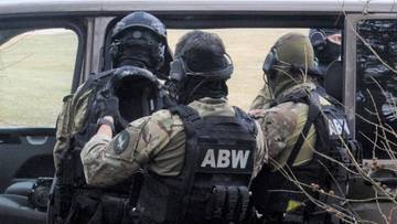 Trzech funkcjonariuszy Agencji Bezpieczeństwa Wewnętrznego w pełnym umundurowaniu i wyposażeniu taktycznym, z widocznymi kamizelkami z napisem "ABW". Jeden z nich stoi tyłem, dwaj pozostali są odwróceni bokiem, rozmawiają ze sobą.