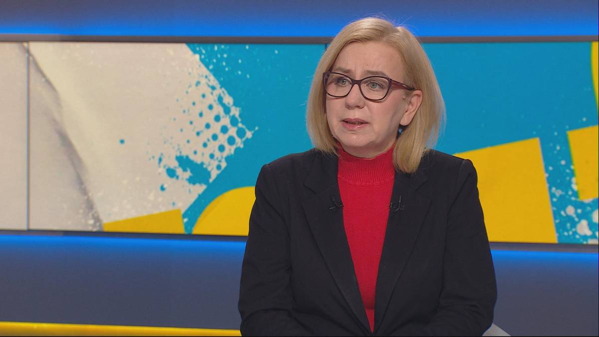 Kobieta w okularach i czarnym żakiecie na tle kolorowej ściany studia telewizyjnego.