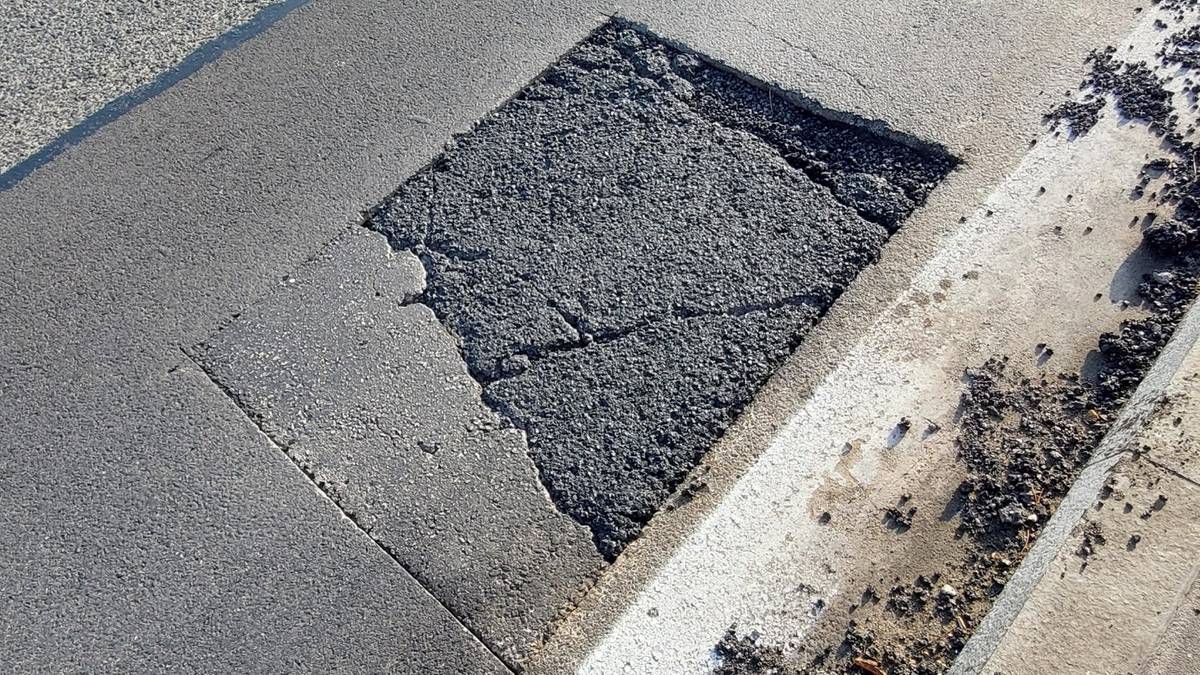 Fragment asfaltowej nawierzchni drogi z widoczną dużą, popękaną wyrwą otoczoną uszkodzonym chodnikiem.