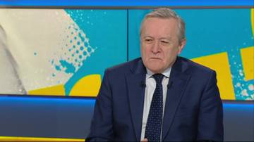 Polityk w garniturze siedzi na tle telewizyjnej scenografii.