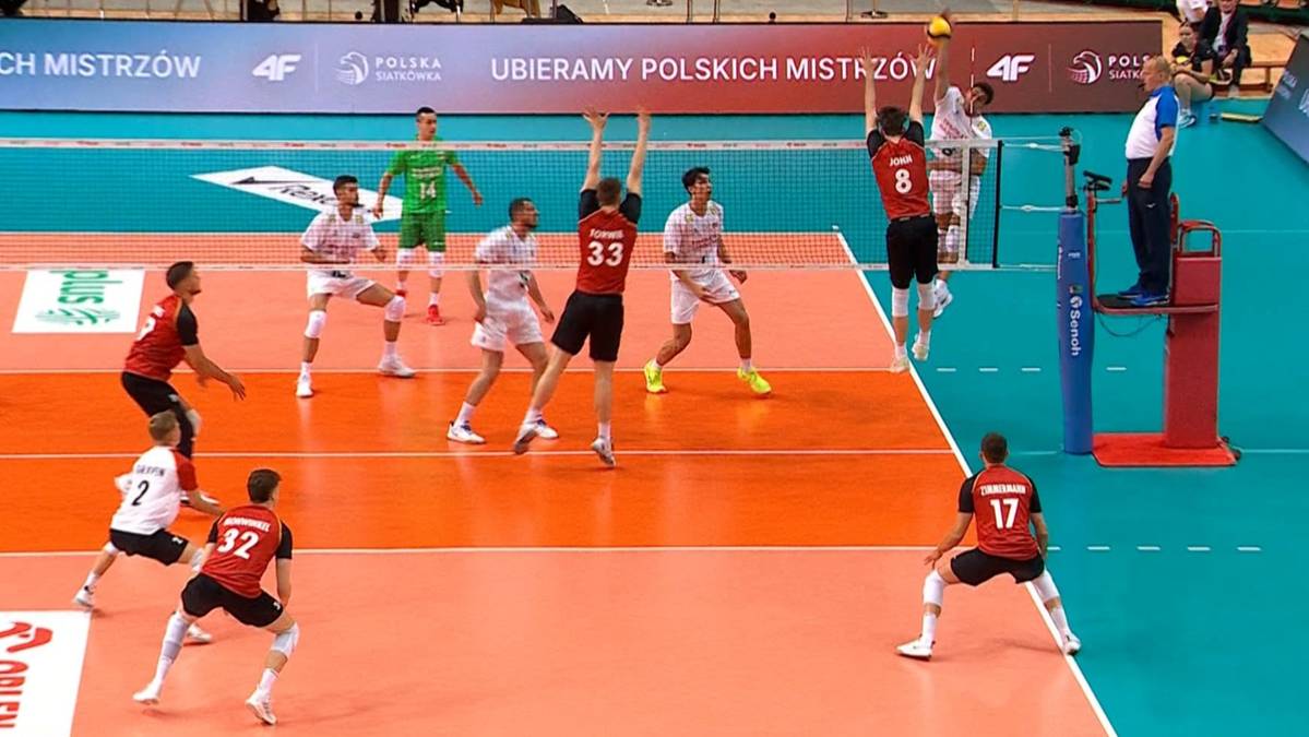 Siatkarze rozgrywają piłkę w meczu, jeden zawodnik wykonuje zagrywkę z wyskoku, pozostali są w gotowości do odbioru lub ataku.