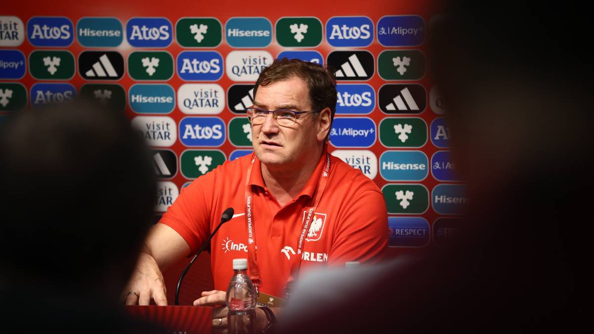 Michał Probierz w czerwonej koszulce reprezentacji Polski siedzi przy mównicy z mikrofonem i butelką wody.