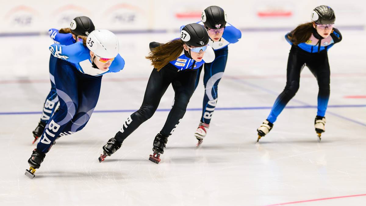 Cztery osoby na łyżwach szybkich ścigają się na torze, wszystkie w kaskach i sportowych strojach.