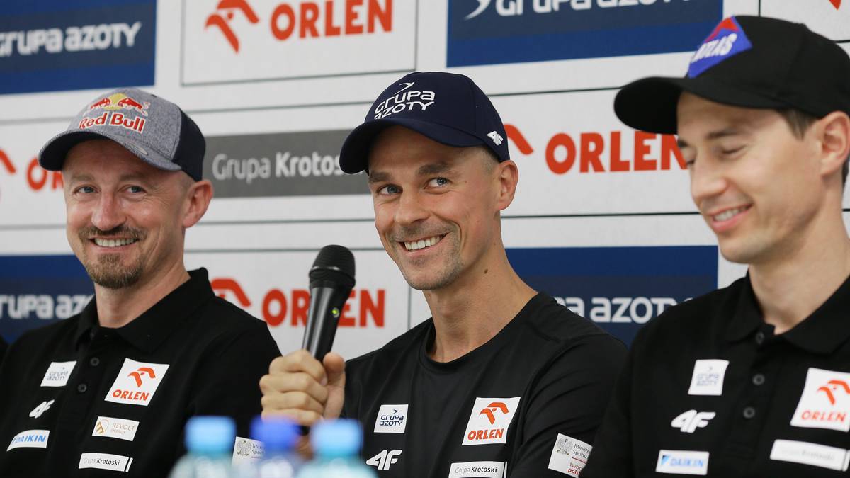 Trzech mężczyzn, prawdopodobnie skoczk&oacute;w narciarskich, podczas konferencji prasowej, jeden z nich trzyma mikrofon.