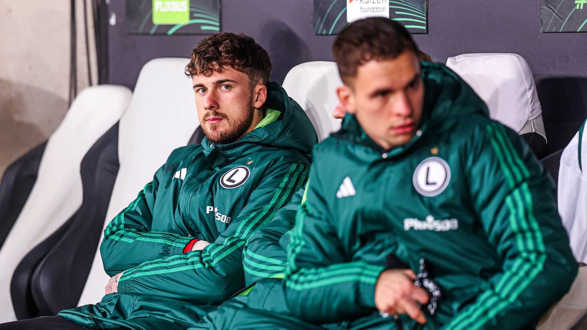 Dw&oacute;ch piłkarzy Legii Warszawa w zielonych kurtkach siedzi na ławce rezerwowych.