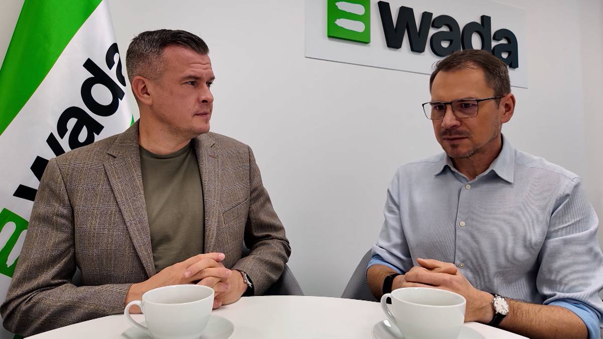 Dwóch mężczyzn siedzi przy stole, rozmawiając. Na ścianie za nimi widnieje logo WADA i flaga z logo WADA.