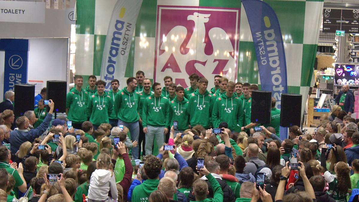 Siatkarze Indykpolu AZS Olsztyn w zielonych strojach stoją na scenie przed grupą kibiców, z banerami sponsorów i herbem klubu w tle.