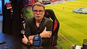 Mężczyzna w okularach siedzi za stołem, z rękami skrzyżowanymi na piersi. Przed nim stoi mikrofon z logiem Polsat Sport. W tle widać stadion.