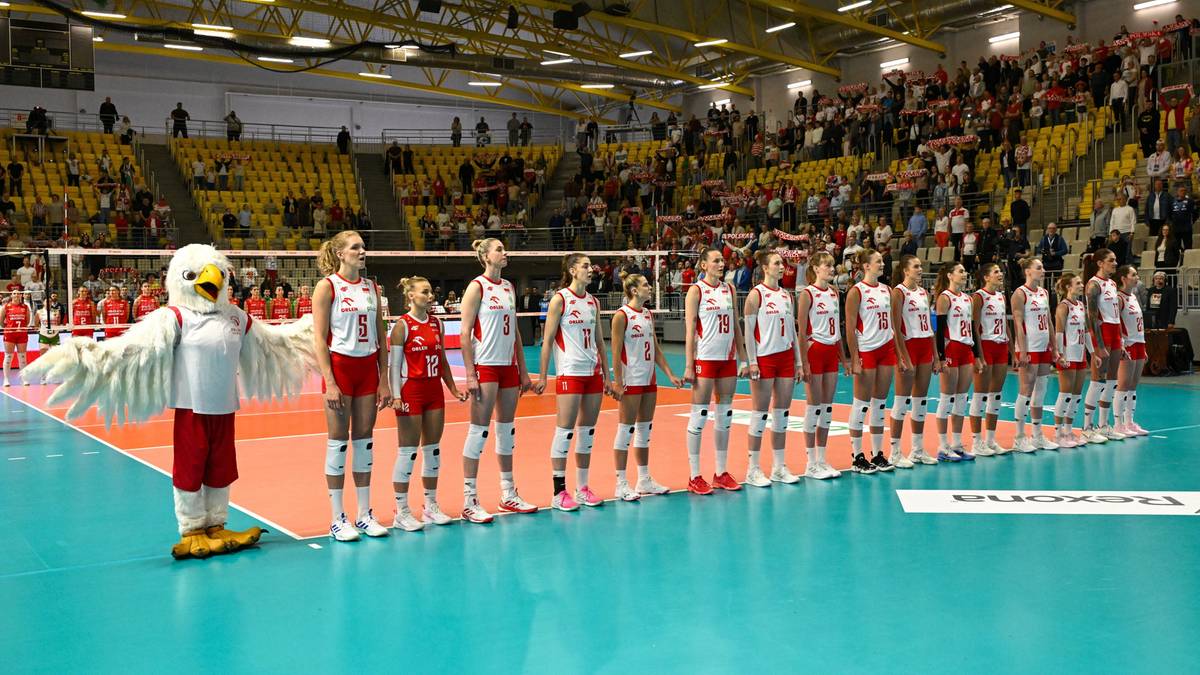 Drużyna siatkarek ustawiona na boisku w białych i czerwonych strojach, z maskotką sokoła po lewej stronie. Widownia na trybunach.