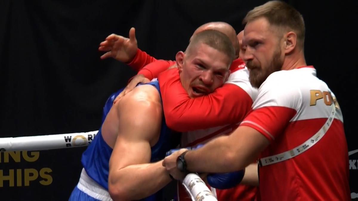 Dwóch bokserów w narożniku. Jeden w niebieskim stroju jest przytrzymywany przez drugiego w czerwono-białym stroju, który prawdopodobnie jest jego trenerem.