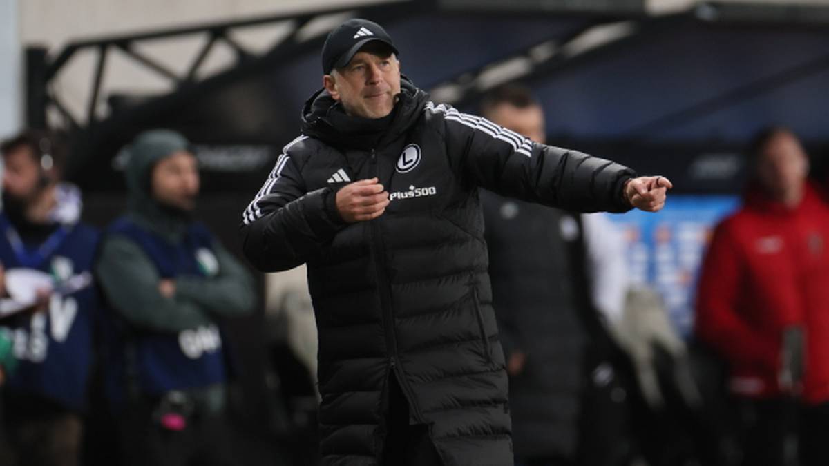 Trener w czarnej kurtce i czapce z daszkiem wskazuje palcem w bok.