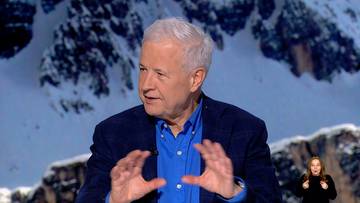 Starszy mężczyzna z siwymi włosami w granatowej marynarce i niebieskiej koszuli, gestykuluje rękami przed aparatem. W tle widoczne są zaśnieżone g&oacute;ry.