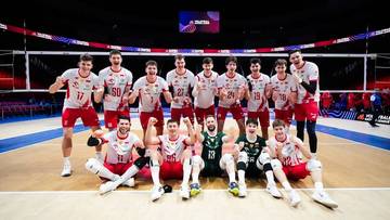 Drużyna polskich siatkarzy w białych strojach sportowych z czerwonymi elementami pozuje do zdjęcia na boisku siatkarskim.