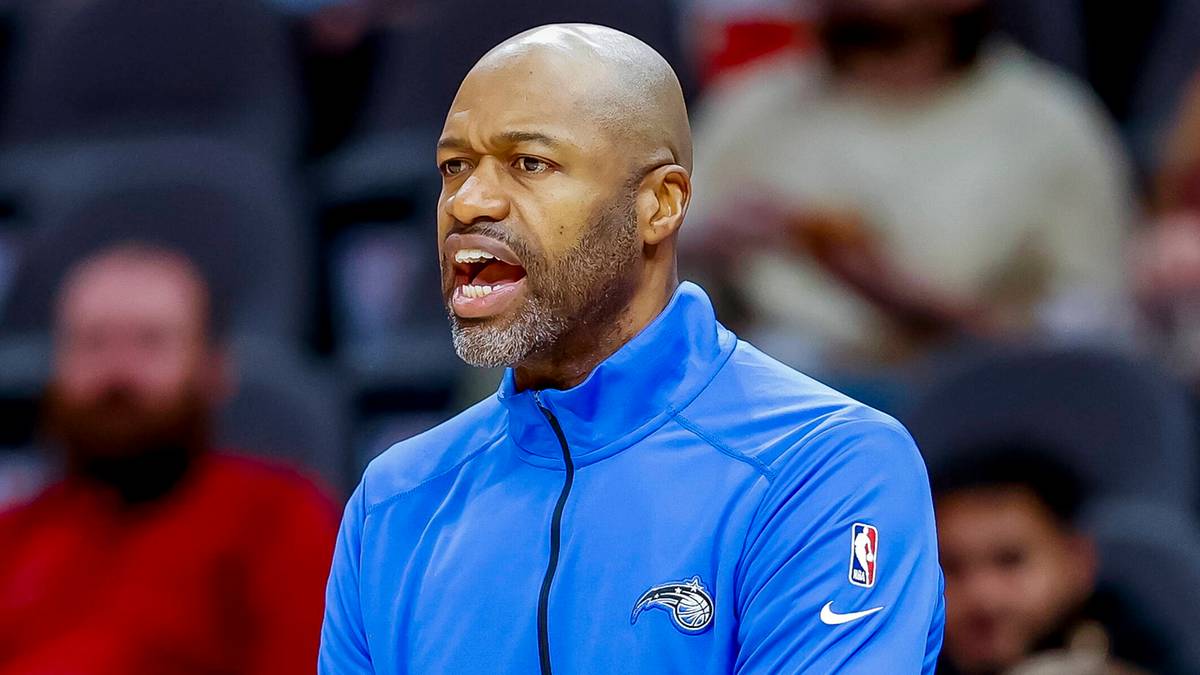 Mężczyzna w niebieskiej kurtce z logo Orlando Magic, ubrany w str&oacute;j sportowy, przemawia z rękami skrzyżowanymi na piersi.