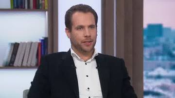 Mężczyzna w czarnym garniturze i białej koszuli siedzi na krześle, na tle widać regał z książkami i okno z widokiem na miasto.