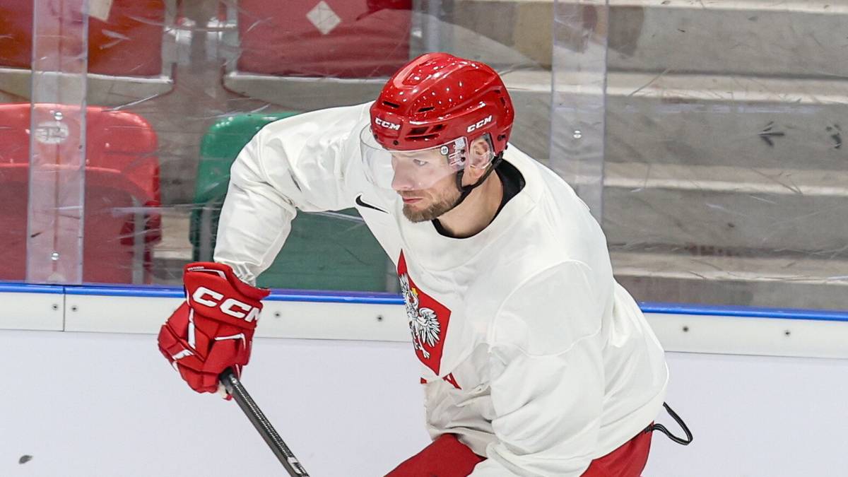 Hokeista w białym stroju z czerwonymi elementami, w czerwonym kasku, z kijem hokejowym na lodzie.