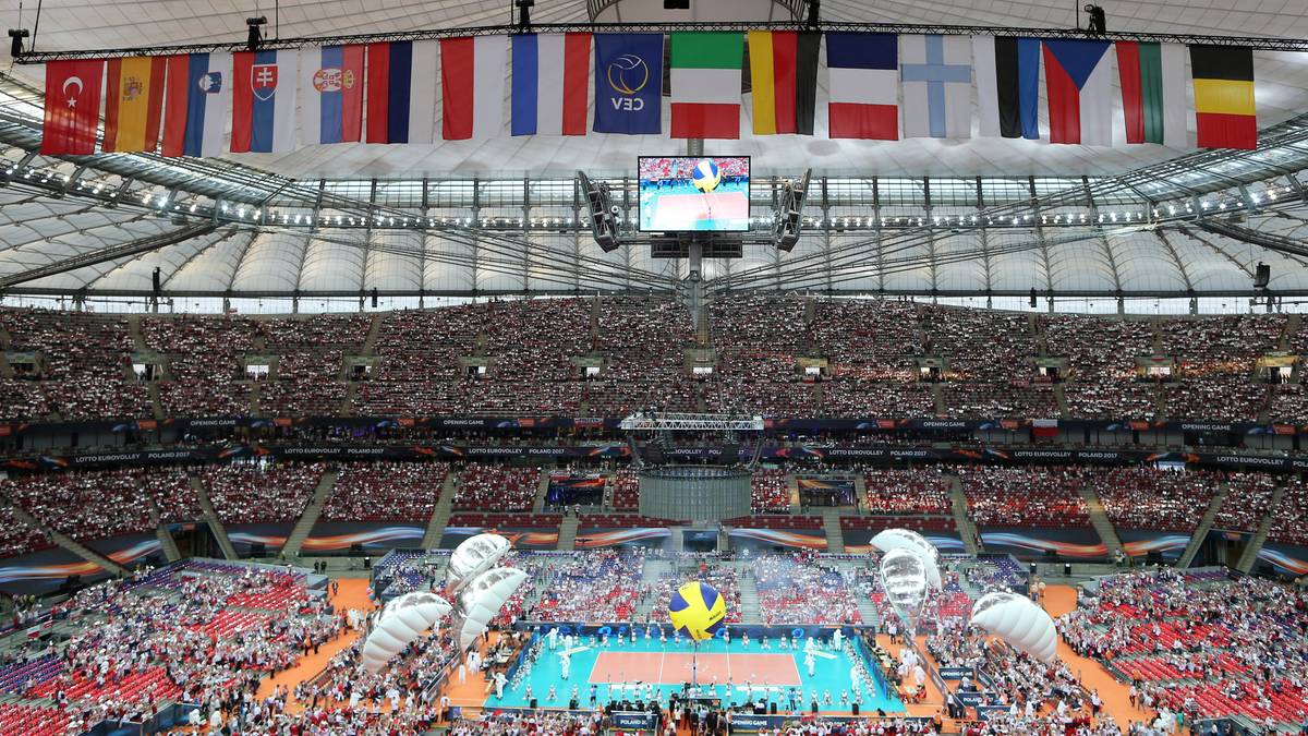 Widok stadionu siatkarskiego z trybunami wypełnionymi publicznością, boiskiem i flagami państwowymi nad nim.