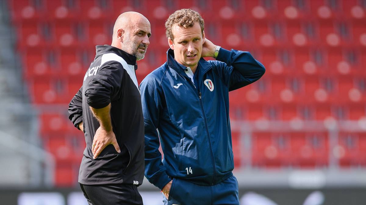 Dwóch mężczyzn w strojach sportowych stoi na boisku piłkarskim, na tle czerwonych trybun.