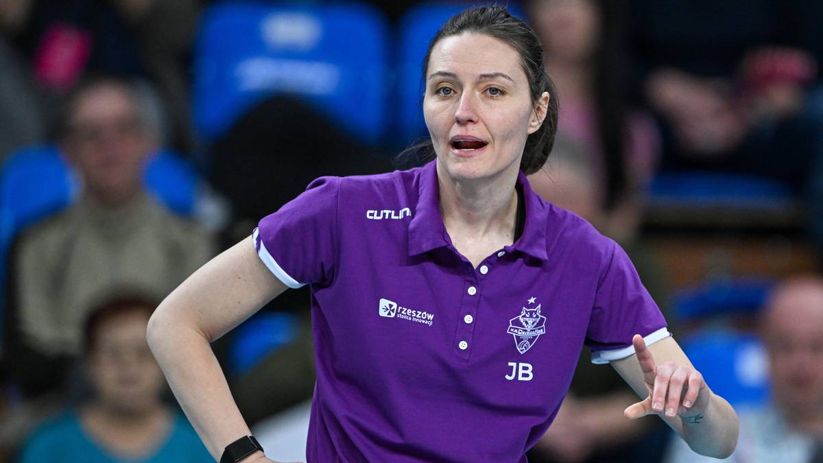 Kobieta w fioletowej koszulce sportowej z inicjałami JB, wydająca polecenia na tle trybun.