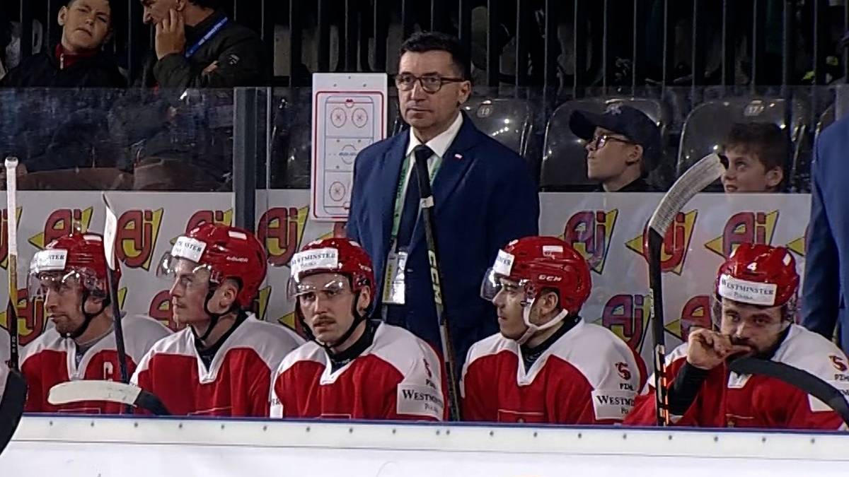 Drużyna hokeist&oacute;w w czerwono-białych strojach i kaskach siedzi na ławce rezerwowych. W tle stoi trener.