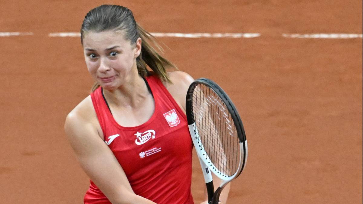 Tenisistka Maja Chwalińska w czerwonym stroju sportowym z biało-czerwoną flagą Polski na piersi, z rakietą tenisową w dłoni, podczas meczu na ceglanym korcie.