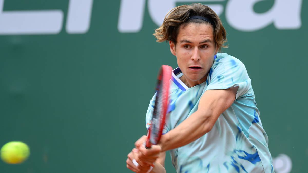 Tenisista z jasnymi włosami, ubrany w jasnoniebieską koszulkę z wzorem, uderza rakietą czerwoną piłkę tenisową podczas meczu.