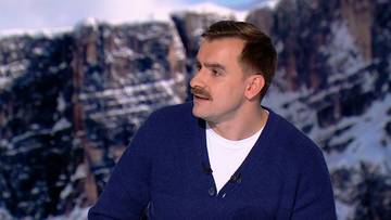 Mężczyzna w średnim wieku, ubrany w granatowy sweter i białą koszulkę, patrzy w bok z lekko otwartymi ustami. W tle widoczne są ośnieżone g&oacute;ry.