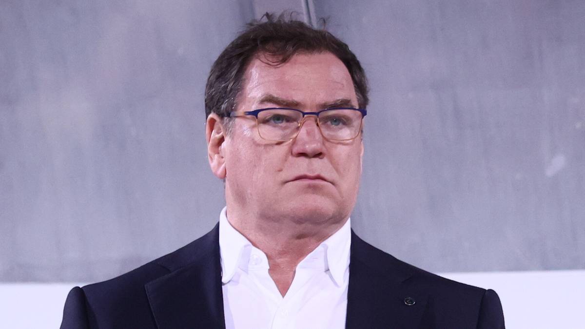 Mężczyzna w garniturze i okularach stoi na tle trybun stadionu piłkarskiego.