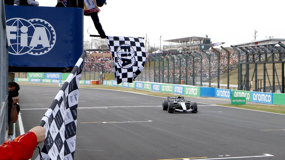 Samoch&oacute;d Formuły 1 na torze wyścigowym zbliża się do mety, gdzie powiewają flagi w szachownicę.